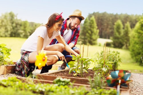 A young woman and a young man are working together in a garden with neatly arranged raised wooden beds containing various green plants and young shrubs. The woman, wearing a white top, plaid shorts, and a headband, is crouching down and tending to the plants, while the man, dressed in a casual shirt, jeans, and a straw hat, is also engaged in gardening tasks. The garden area features well-maintained, lush grass surrounding the beds, and the background shows a natural landscape of trees and an open field under a clear sky with natural daylight. Gardening tools and colorful decorative garden ornaments are visible in the foreground, indicating outdoor maintenance and planting activities. This scene reflects a well-organized outdoor space that could be part of a private garden or community green area, suitable for professional gardening and landscaping services such as those offered by Lawn Mowing Blackfriars in the Blackfriars area of London, near postcode EC4, emphasizing outdoor enjoyment and active gardening in a scenic environment.